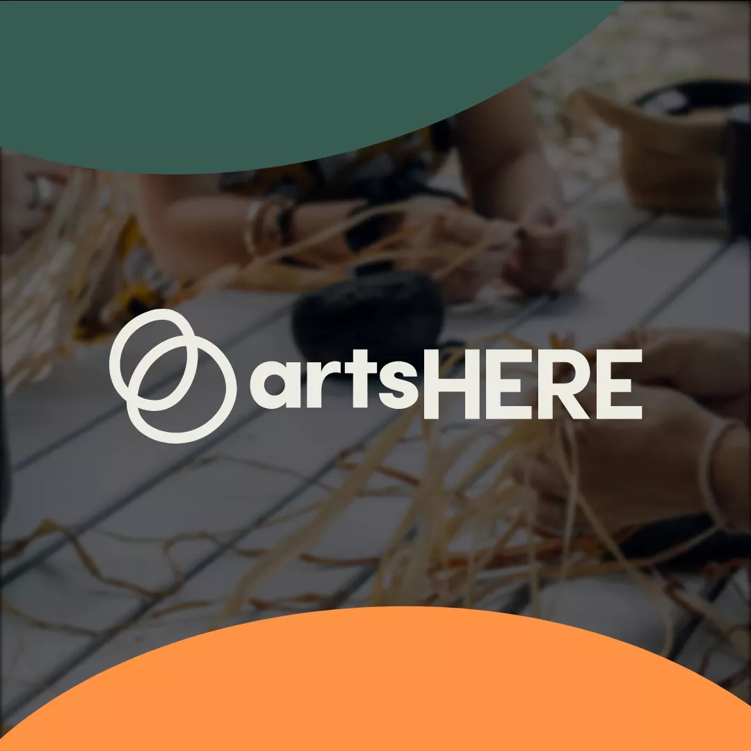 The artsHERE logo in white features two overlapping spheres. It’s framed by the edge of two spheres, one green and one orange. The logo is over a blurred image of light-skinned women weaving hay at a picnic table.